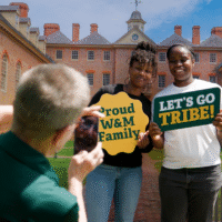 A person takes a photo of two people holding signs in front of the Wren Building.