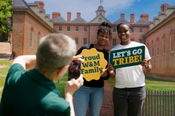 A person takes a photo of two people holding signs in front of the Wren Building.