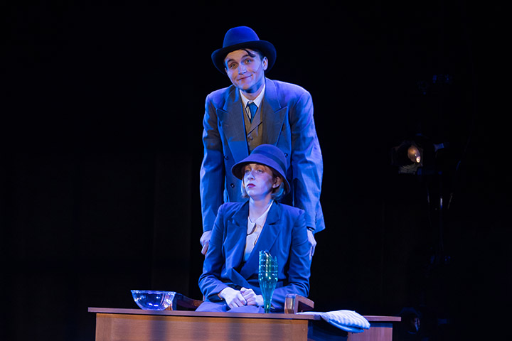 A person stand behind another person, sitting down, on a stage.