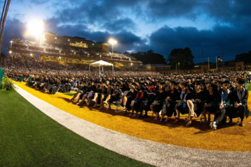 Graduates in caps and gowns sit in a large football stadium.