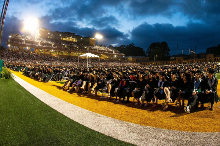 Graduates in caps and gowns sit in a large football stadium.