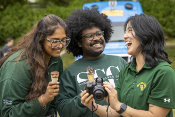 Two people laugh while eating ice cream cones while a third person shows them a camera.