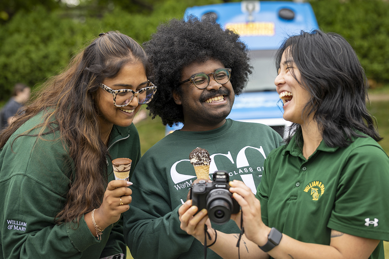 Two people laugh while eating ice cream cones while a third person shows them a camera.