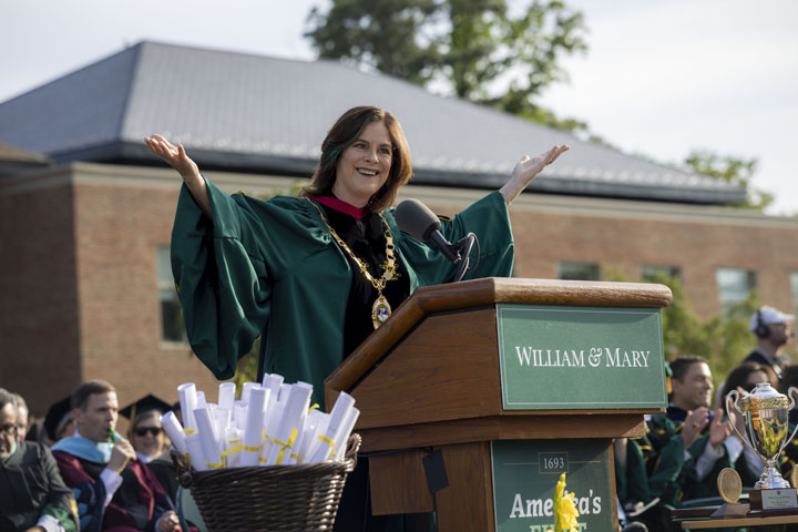 Katherine Rowe raises both arms and smiles with standing at a podium with a William & Mary sign on it.
