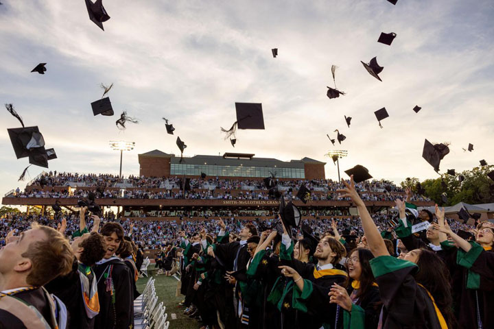 People in academic gowns throw their caps in the air inside a football stadium.