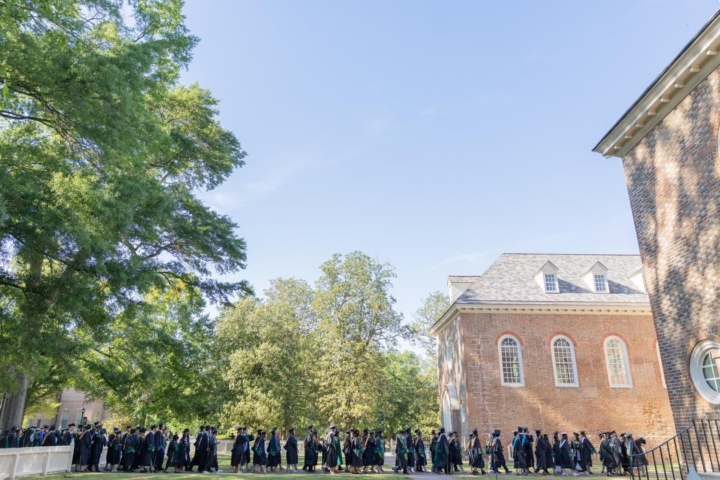 A line of people in caps and gowns leads into the Wren Building.