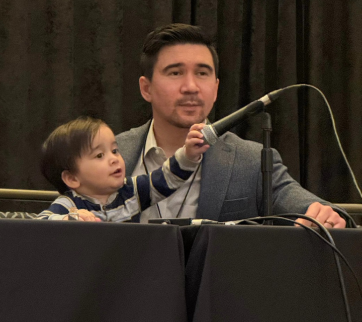 A child reaches for a microphone while sitting on his father's lap at a table.