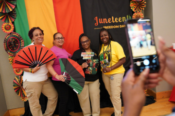 Four people pose with prop in front of a Juneteenth backdrop while someone else takes a photo.