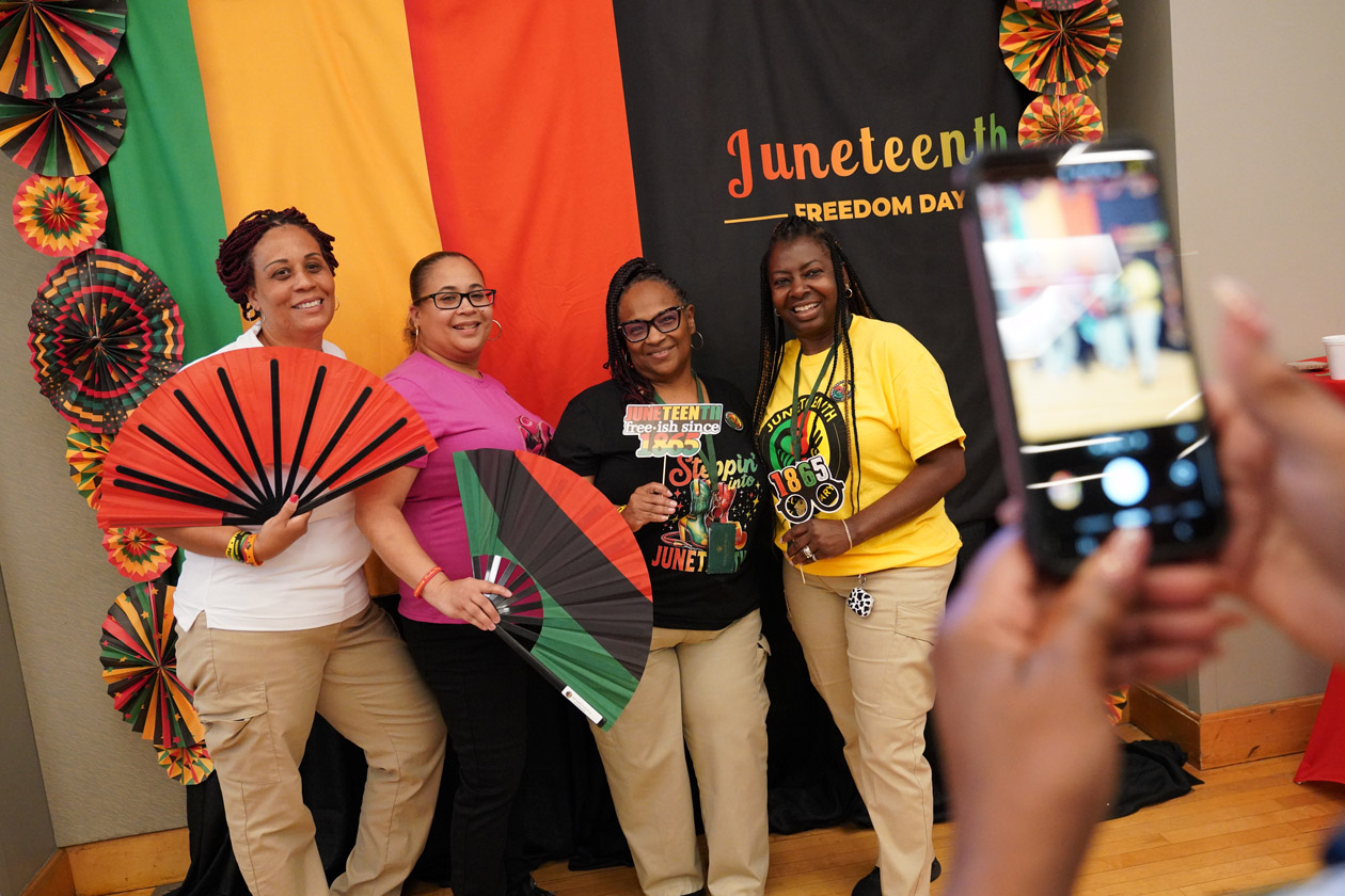 Four people pose with prop in front of a Juneteenth backdrop while someone else takes a photo.