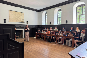 A person stands toward the left of the image with a microphone in hand, speaking to two rows of young students. On the left side of the wall is a map of the thirteen colonies.