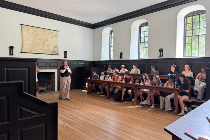 A person stands toward the left of the image with a microphone in hand, speaking to two rows of young students. On the left side of the wall is a map of the thirteen colonies.