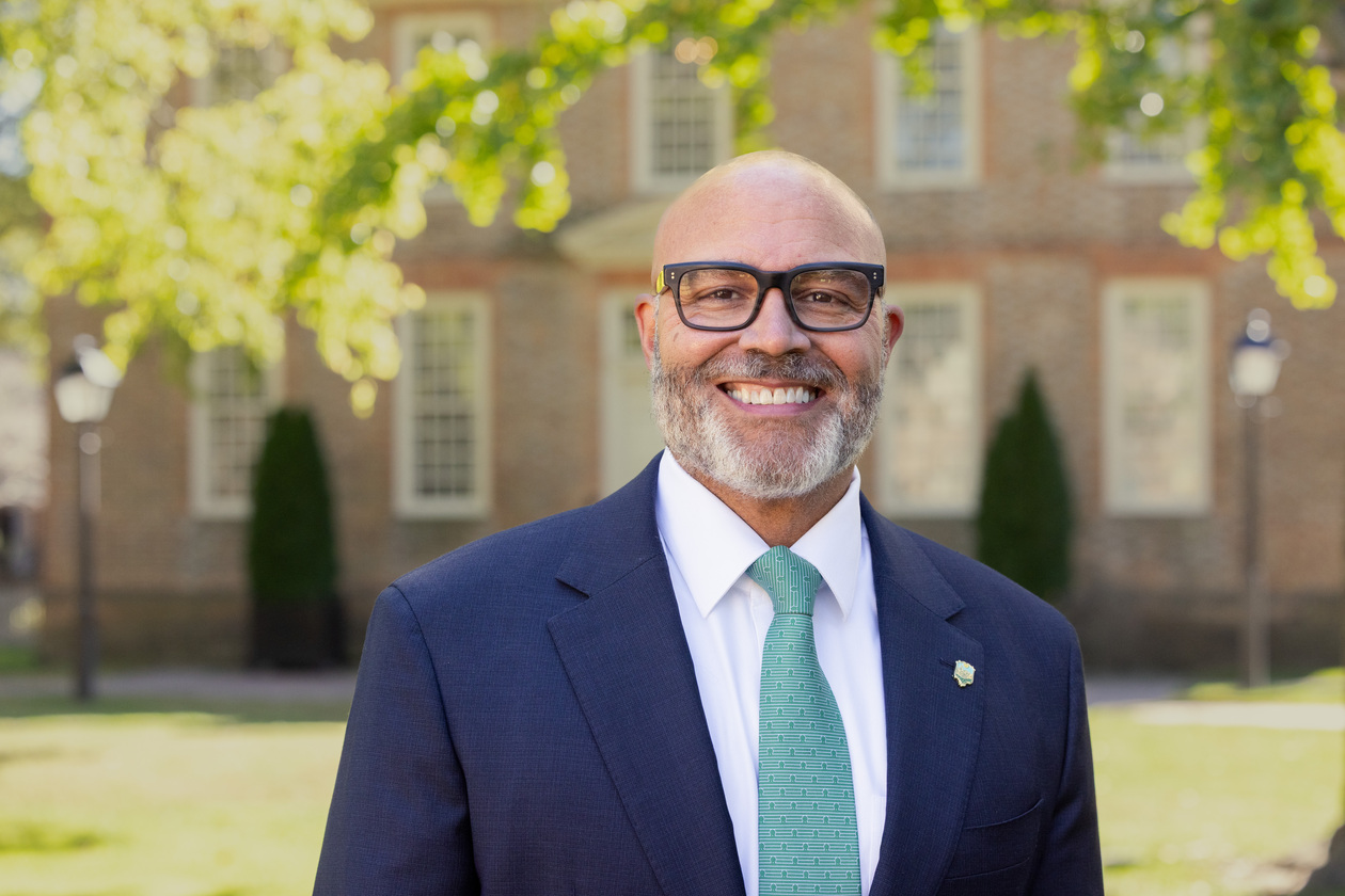 Michael Powell stands in front of the Wren Building. He is smiling and wearing glasses, a blue blazer and a green tie.