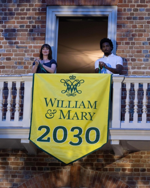 Two students stand on a white balcony with a yellow "William & Mary 2030" banner hanging over it.