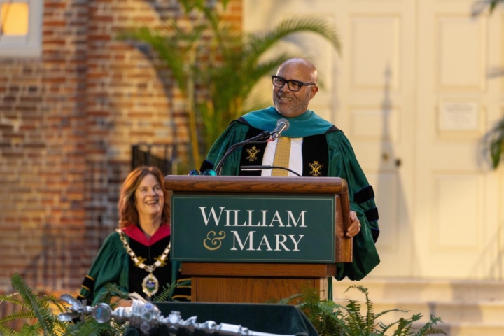 Convocation Speaker Michael K. Powell wears a green robe and stands behind a podium. President Katharine Rowe sits behind him.