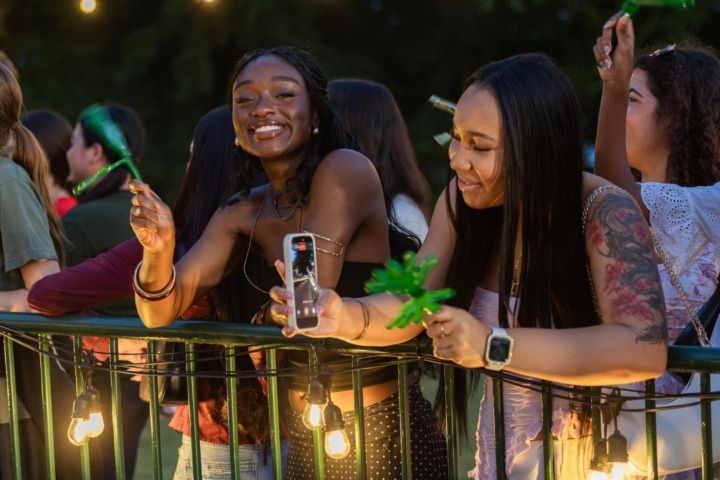 A student smiles facing the camera while holding clappers. Another is angled toward the Wren Building recording a video on their phone.