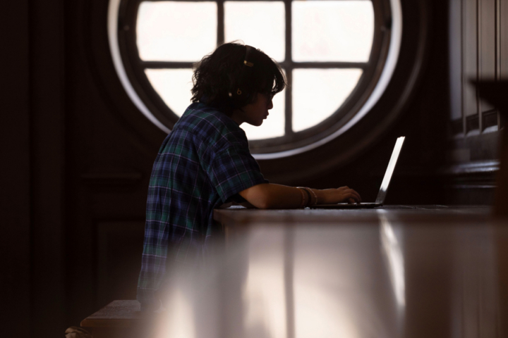 A student works on their computer in the Wren Building. We only see their side profile and light from a large circular window casts light in the photo.