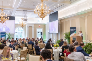 People sit at tables in a large room and listen to a person as a podium.