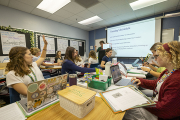 People sit around tables in a classroom and listen to a teacher.