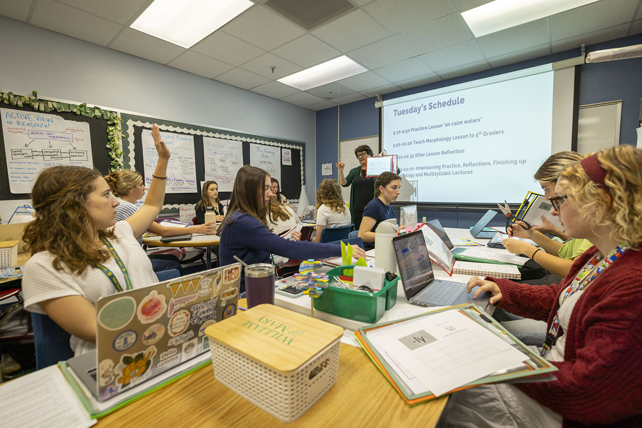 People sit around tables in a classroom and listen to a teacher.