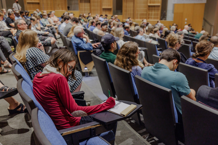 People sit in chairs in an auditorium and take notes.