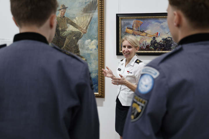 A person in a military uniform speaks while standing next to pieces of art on a wall.