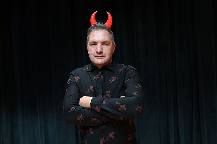 Andrew Fisher wears fake devil's horns on his head and crosses his arms.