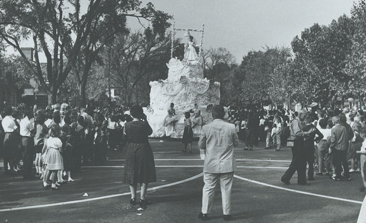 A black and white photos shows a parade float shaped like a large wedding cake,