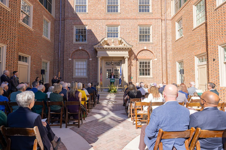 People sit in chairs in an outdoor courtyard while a person speaks at a podium.