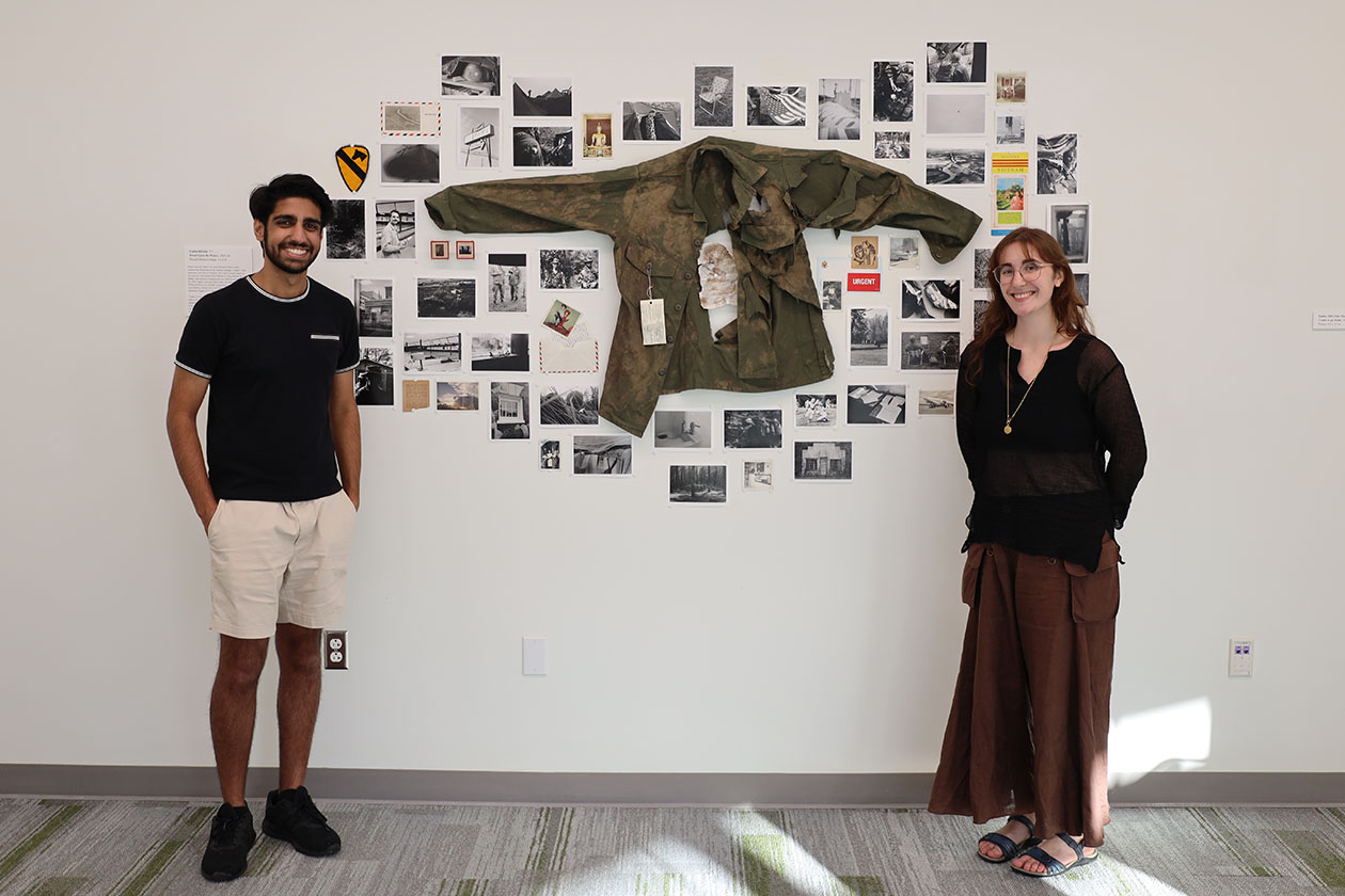 Two people pose for a photo near a display on a wall including photos and a jacket.