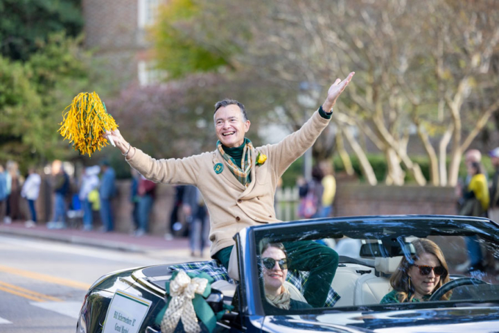 A person ride on the back of a convertible and waves a pom-pom.