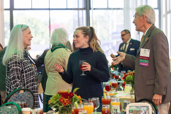 People chat with one another while standing next to a table with food on it.