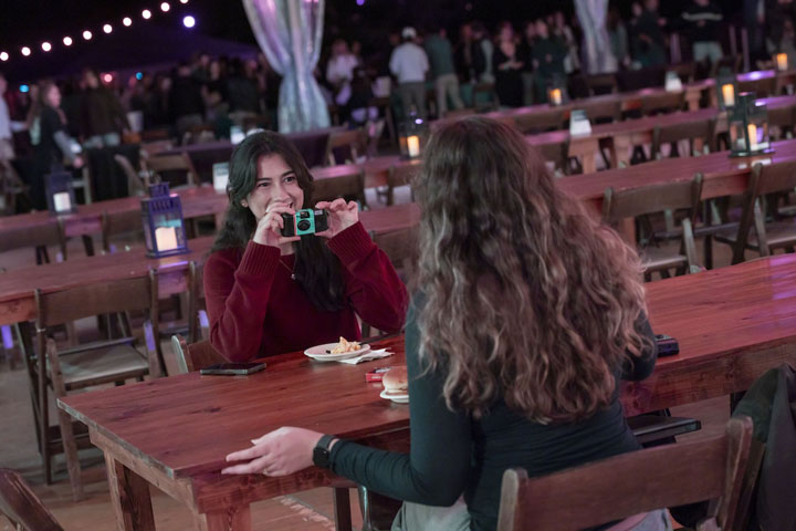 A person sitting at a table takes a photo of another person with a disposable camera.